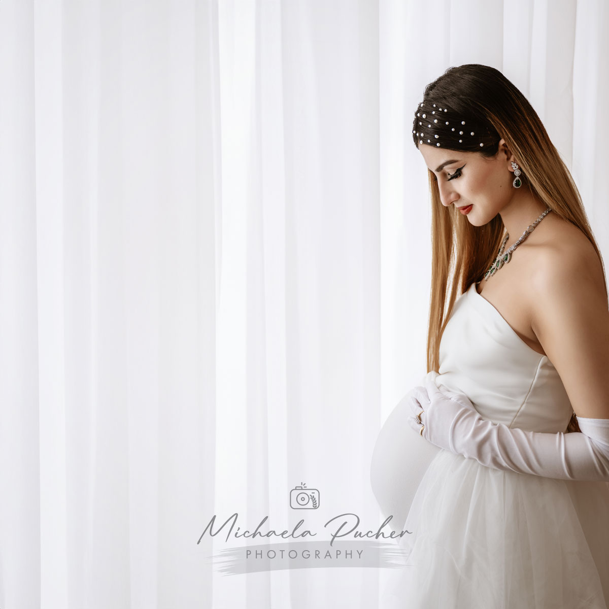 Elegante schwangere Frau in weißem Kleid und Perlen-Haarband steht seitlich am Fenster und hält ihren Babybauch.